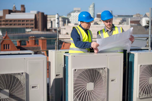 Two HVAC workers reviewing blueprints while installing rooftop units, demonstrating teamwork and technical skills.